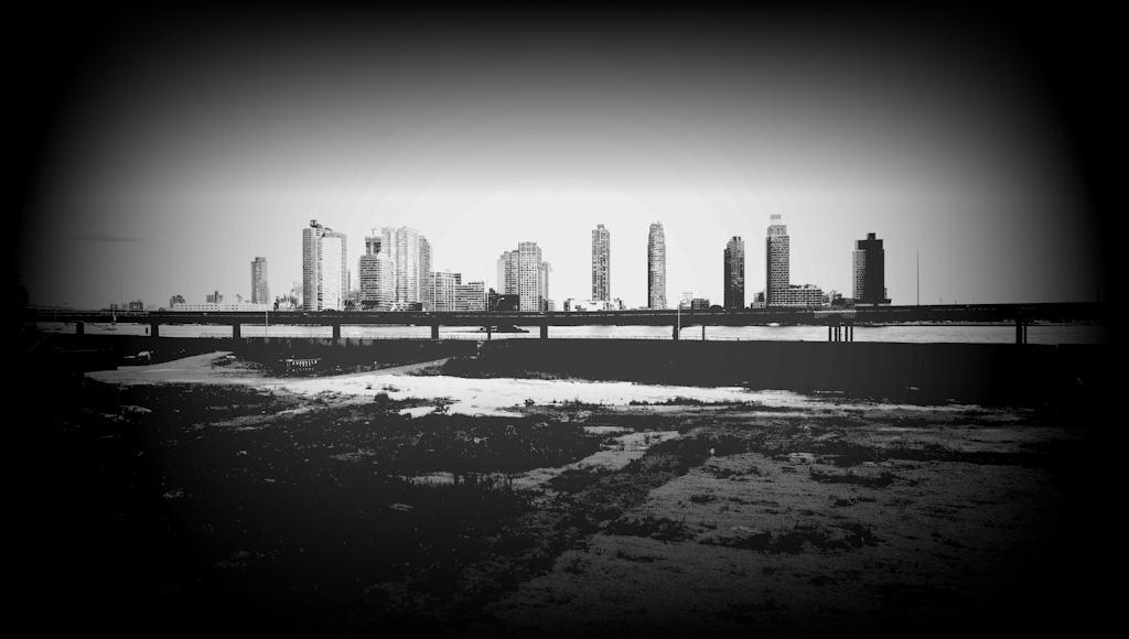 A picture of a large empty plot of land beside a river with a populous city skyline in the background.